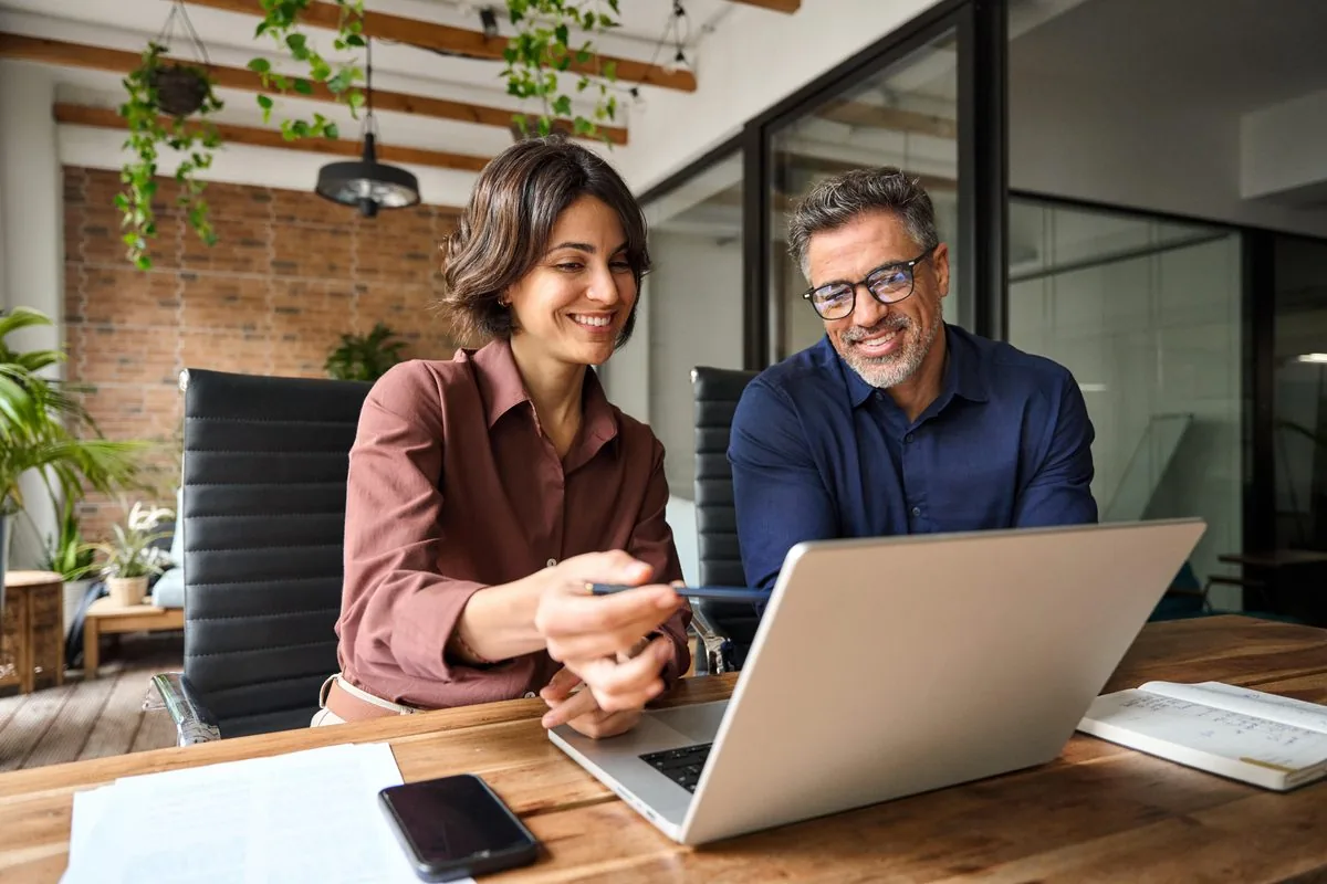 Two colleagues smiling and reviewing work together on a laptop in a modern office