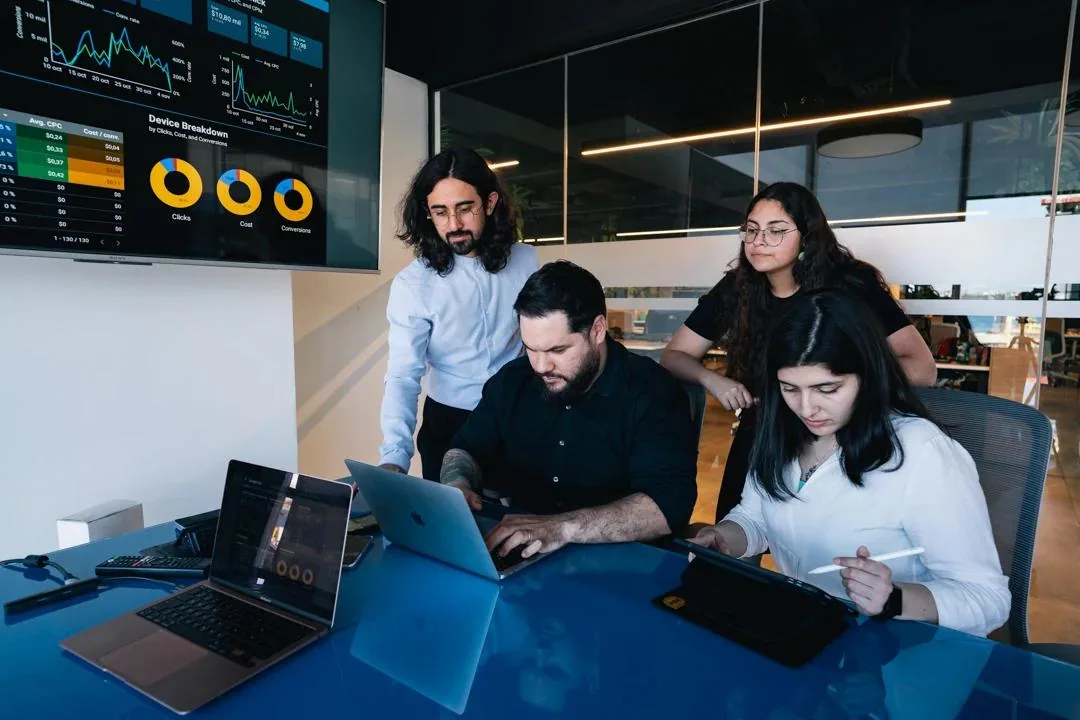 Team members gathered around a table looking at laptops, with an analytics dashboard displayed on a screen behind them